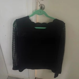 Svart blus med transparenta ärmar - Säljer en svart blus med unika transparenta ärmar i nätmaterial och vågig kant vid halsen. Perfekt för dig som vill ha något stilrent men ändå med en twist. i storlek XL men kan definitivt användas som M/L