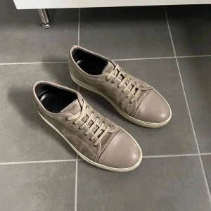 Beige sneakers i mocka och skinn från Lanvin - Säljer ett par stilrena beige sneakers från Lanvin med ovandel i mocka och tåparti i skinn. Skorna har snörning och en platt vit sula. Perfekta för dig som gillar en klassisk och clean look.
