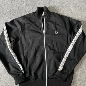 Svart track jacket från Fred Perry - Säljer en svart track jacket från Fred Perry med vita detaljer och logga på bröstet. Jackan har dragkedja framtill och vita band med Fred Perry-loggor längs ärmarna. Perfekt för en sportig och stilren look. Nästan aldrig använd. 