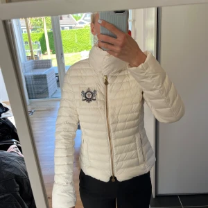 Vit dunjacka från Morris Lady - Snygg vit dunjacka från Morris Lady med broderad logga på bröstet och guldfärgad dragkedja. Jackan har hög krage, figurnära passform och två fickor framtill. Perfekt för kyligare dagar.