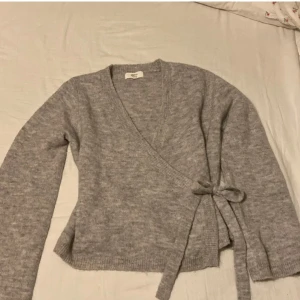 Grå omlottkofta från Gina Tricot - Mysig grå omlottkofta från Gina Tricot i ullblandning. Tröjan har långa ärmar, knyt i sidan och ribbade kanter. Perfekt för lager-på-lager och enkel att styla till olika outfits.
