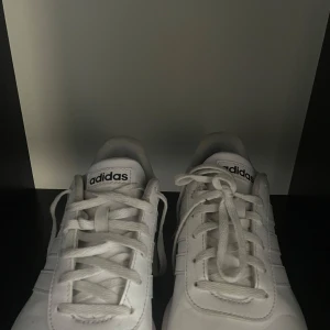 Äkta Vita sneakers från Adidas - Säljer ett par klassiska vita sneakers från Adidas med snörning och diskret logga på hälen och plösen. Skorna har en platt sula och är tillverkade i syntetmaterial. Perfekta för en stilren och sportig look.