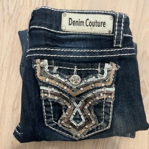 Miss Me Inspirerade Jeans - Snygga mörkblå jeans från Denim Couture med unika broderade och dekorerade bakfickor i silver och brons. Jeansen har kontrastsömmar och klassisk femficksdesign. Perfekta för dig som vill sticka ut med detaljer! Längd: 96cm midja: 35 stretch till 38-40cm. Benöppning: 21cm