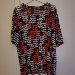 Mönstrad t-shirt i svart, rött och vitt - Säljer en cool t-shirt med grafiskt mönster i svart, rött, vitt och rosa. Tröjan har normal passform och korta ärmar. Perfekt för dig som vill sticka ut lite extra med din stil! Ett linne under blusen gör den skit snygg! 