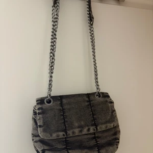 Grå axelväska med kedjerem - Säljer en snygg grå axelväska med kedjerem och rutigt mönster. Väskan har lockstängning och ett innerfack med dragkedja. Perfekt för dig som vill ha en stilren accessoar med lite edge.köppt gör 600 kr. Aldrig använd( pris går diskuter)