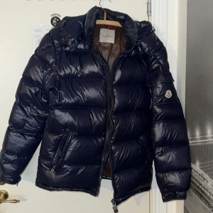 Blå pufferjacka från Moncler - Säljer en snygg blå pufferjacka från Moncler. Jackan har en glansig finish med dragkedja och huva. Perfekt för kyliga dagar när du vill hålla stilen. Moncler-logga på ärmen ger en exklusiv touch.