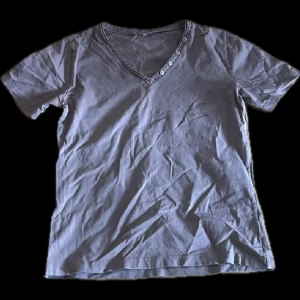 Brun t-shirt med v-ringning och knappar - Säljer en stilren grå t-shirt med v-ringning och dekorativa knappar längs halsringningen. Perfekt för en avslappnad look. Passar bra till jeans eller shorts.