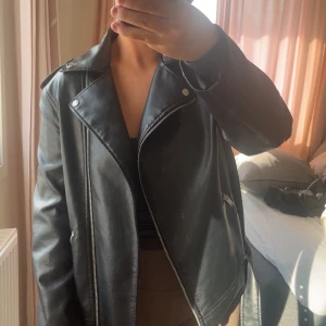 Svart skinnjacka med dragkedja - Snygg svart skinnjacka med dragkedja och klassisk bikerstil. Jackan har långa ärmar och detaljer som knappar och dragkedjor på framsidan. Perfekt för en cool och stilren look.