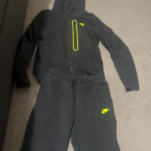 Nike tech - Säljer ett par svarta mjukisbyxor från Nike med en gul Nike-logga på benet. Byxorna har snörning i midjan för justerbar passform. Perfekta för en avslappnad stil.