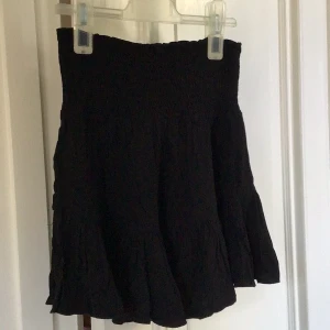 Svart kortkjol med volang - Säljer en svart kortkjol med volangdetaljer. Kjolen har en elastisk midja som ger en bekväm passform. Perfekt för att skapa en stilren look.