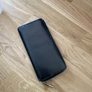 Svart plånbok med dragkedja - Säljer en stilren svart plånbok i skinn med dragkedja runtom. Plånboken har flera fack för kort och en separat sektion med dragkedja för mynt. Från märket Saddler.