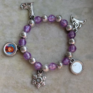 Lila och silver armband med berlocker - Charmigt armband med lila och silverfärgade pärlor. Utsmyckat med berlocker, inklusive en snöflinga och en karaktärsbild. Perfekt för att lägga till en lekfull touch till din stil.