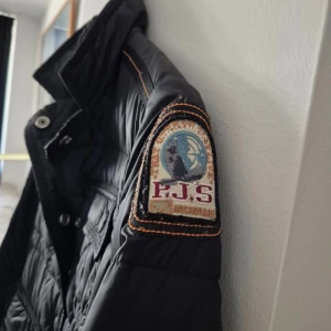 Svart dunjacka från Parajumpers - Säljer en svart dunjacka från Parajumpers med quiltad design och märkespatch på ärmen. Jackan har en lättviktskonstruktion och stängs med knappar och dragkedja. Perfekt för kyliga dagar!