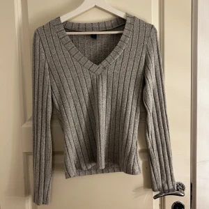 Grå ribbad tröja med v-ringning - Säljer en stilren grå ribbad tröja med v-ringning. Tröjan har långa ärmar och är perfekt för kyligare dagar. Passar bra till både jeans och kjol.