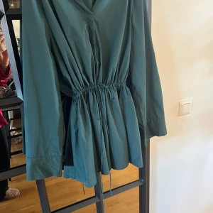 Grön blus skjorta vet inte  från Lindex - Säljer en stilren grön skjorta från Lindex med knytning i midjan. Skjortan har långa ärmar och en lös passform, perfekt för vår och höst. Den är tillverkad i ett lätt material som gör den bekväm att bära.