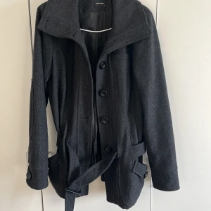 Grå kappa från Vero Moda - Snygg grå kappa från Vero Moda med knappar och bälte i midjan. Perfekt för kyligare dagar med sin stilrena design och praktiska fickor. Kappan har långa ärmar och en klassisk krage.