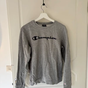 Grå sweatshirt från Champion - Säljer en klassisk grå sweatshirt från Champion med deras ikoniska logga i svart på bröstet. Tröjan har långa ärmar och en rund halsringning. Obs. Storlekarna är för barn, för en ungdom passar den en person med S.