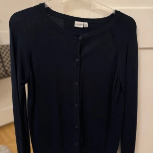 Marinblå kofta från Basic Needs - Säljer en stilren marinblå kofta från Basic Needs. Koftan har långa ärmar och knäpps med knappar framtill. Perfekt för lager-på-lager och passar till många olika outfits.