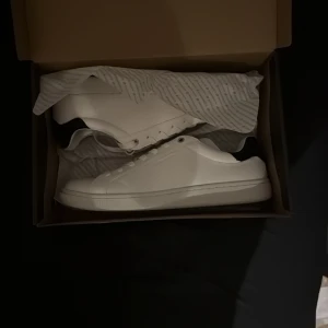 Vita sneakers från Björn Borg - Säljer ett par stilrena vita sneakers från Björn Borg. Skorna har en klassisk design med snörning och är perfekta för en avslappnad look. De är i storlek EU 45 och kommer i originalförpackning. Aldrig använda behöver pengarna för ett annat köp!