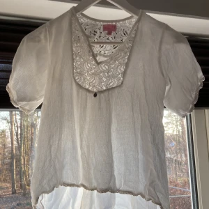 Vit blus med spetsdetaljer från Stories - Säljer en söt vit blus från Stories med vackra spetsdetaljer vid halsringningen. Blusen har korta ärmar och en luftig passform, perfekt för varmare dagar. Den har en lätt transparent look och en elastisk nederkant för en bekväm passform.