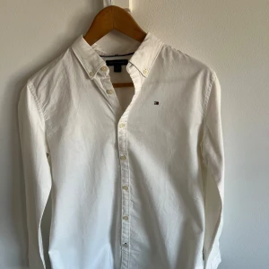 Vit skjorta från Tommy Hilfiger - Säljer en klassisk vit skjorta från Tommy Hilfiger med button-down krage och broderad logotyp på bröstet. Skjortan har långa ärmar och knappar framtill. Perfekt för en stilren look.