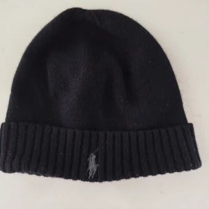 Svart stickad mössa från Ralph Lauren - Snygg svart stickad mössa från Ralph Lauren med ribbad kant och broderad logga framtill. Perfekt för kyliga dagar.
