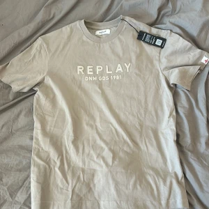 Beige t-shirt från Replay - Säljer en snygg beige t-shirt från Replay med texten 'REPLAY DNM GDS 1981' tryckt på framsidan. T-shirten är kortärmad och har en klassisk rund hals. Perfekt för en avslappnad stil.