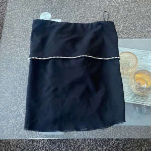 Svart axelbandslös top med pärldetalj - Elegant svart axelbandslös top med en fin pärldetalj som löper horisontellt över bröstet. Perfekt för en stilren och sofistikerad look.