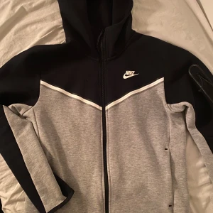 Grå och svart hoodie från Nike - Säljer en snygg grå och svart hoodie från Nike med dragkedja och huva. Jackan har en vit Nike-logga på bröstet och en praktisk ficka med dragkedja på ärmen. Perfekt för en sportig look!(priset kan diskuteras!!!!)