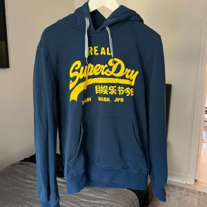 Blå hoodie från Superdry - Säljer en snygg blå hoodie från Superdry med gul text på framsidan. Den har en klassisk känguruficka och justerbar huva med snören. Perfekt för en avslappnad stil.