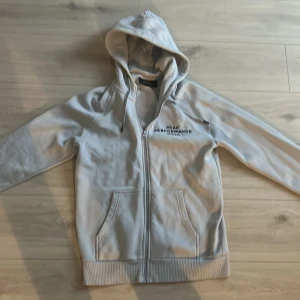Ljus blå hoodie från Peak Performance - Säljer en stilren grå hoodie från Peak Performance med dragkedja och broderad logga på bröstet. Tröjan har en mysig huva och praktiska fickor framtill. Perfekt för kyliga dagar!