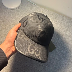 Svart gucci keps - Snygg svart keps från Gucci med det ikoniska GG-mönstret. Kepsen har en justerbar rem baktill för perfekt passform. Tillverkad i ett slitstarkt material med en stilren design som passar till många olika outfits. Storlek ”M”