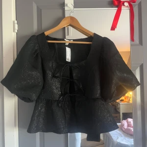 Svart glittrig blus med puffärmar - Snygg svart blus med glittrig yta och puffiga kortärmar. Blusen har en knytning framtill och en lätt croppad passform. Perfekt för en kväll ute eller en festlig tillställning.💕