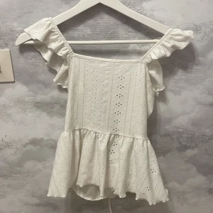 Vit blus från Shein - Säljer en söt vit blus från Shein med volangdetaljer och knytning i ryggen. Blusen har korta ärmar och ett broderat mönster som ger en romantisk känsla. Perfekt för varma dagar!