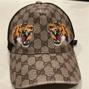 Gucci keps med tigertryck - Snygg keps från Gucci med ikoniskt GG-mönster och två tigrar framtill. Kepsen har en justerbar rem baktill i grönt och rött. Perfekt för att ge din outfit en lyxig touch.
