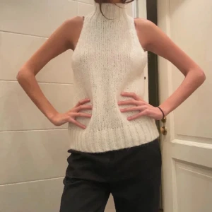 Vit stickad ärmlös polotopp - Säljer en stilren vit stickad polotopp med hög krage. Toppen är ärmlös och har en ribbad nederkant som ger en snygg passform. Perfekt för lager-på-lager eller som den är.