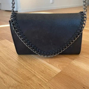 Svart axelväska med kedja - Snygg svart axelväska med kedjedetaljer och flätad kant. Perfekt för att ge din outfit en edgy touch. Väskan har en stilren design och är tillverkad i ett material som liknar mocka. Lite ”skavanker” som syns på bilderna. Nypris 500kr