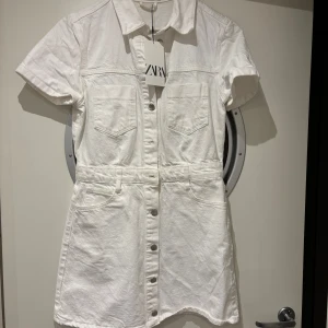 Vit kortärmad jeans klänning från Zara - Säljer en stilren vit klänning från Zara med korta ärmar och knappar framtill. Klänningen har två bröstfickor och en klassisk krage, perfekt för en avslappnad look. Tillverkad i ett bekvämt material som passar bra till varmare dagar. OBS!!! Det är en storlek M men jag har vanligtvis S och den passar på mig!