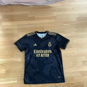 Svart fotbollströja från Adidas real Madrid  - Snygg svart fotbollströja med guldiga detaljer och drakmönster. Tröjan har korta ärmar och klubbmärke på bröstet. Perfekt för fotbollsfans som vill sticka ut på planen.