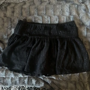 Lågmidjad kjol - Snygg svart lågmidjad kjol med glittrande paljetter. Kjolen har en bred midja och ett lätt utställd passform som ger en fin siluett. Perfekt för att lägga till lite glamour till din outfit.