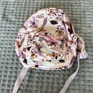 Blommig ryggsäck med rosa och lila nyanser - Säljer en söt ryggsäck med blommigt mönster i rosa och lila nyanser. Ryggsäcken har justerbara axelremmar och en praktisk dragkedja framtill. Perfekt för vardagsbruk eller utflykter.