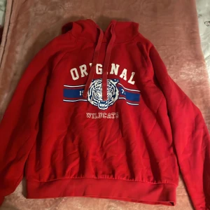 Röd hoodie med tryck - Säljer en röd hoodie med texten 'ORIGINAL WILDCATS' och ett tigertryck i vitt och blått. Hoodien har en klassisk passform med dragsko i huvan och ribbade muddar vid ärmslut och nederkant. Perfekt för en avslappnad stil!