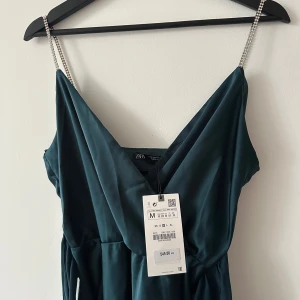 Smaragdgrön ankellång klänning från Zara - Säljer en elegant smaragdgrön ankellång klänning från Zara med smala axelband dekorerade med strass. Klänningen har en omlott-design och en v-ringad halslinje som ger en stilren look. Perfekt för speciella tillfällen.