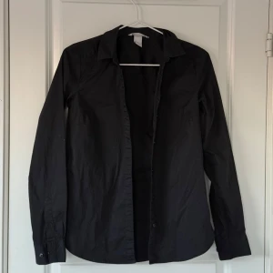 Svart skjorta från H&M - Svart skjorta från H&M i storlek XS. Skjortan har långa ärmar och knappar framtill.