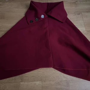 Vinröd cape med dekorativa knappar - Snygg vinröd cape med tre dekorativa knappar i svart och vitt mönster. Perfekt för att ge en elegant touch till din outfit. Capen har en klassisk krage och är tillverkad i ett mjukt material.
