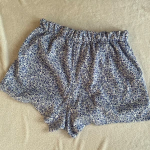 Blå blommiga shorts - Säljer ett par bekväma blå blommiga shorts med elastisk midja. Perfekta för varma sommardagar. De har en lös passform som ger en avslappnad stil.