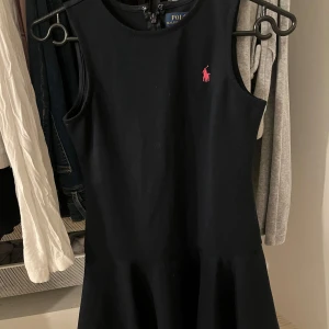Marinblå klänning från Ralph Lauren - Snygg marinblå klänning från Ralph Lauren med en rosa logga på bröstet. Klänningen är ärmlös och har en dragkedja i ryggen för enkel påklädning. Perfekt för en stilren look.