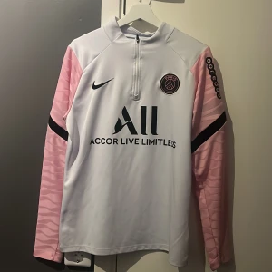 PSG träningströja - Säljer en snygg vit fotbollströja från Nike med rosa ärmar och svart tryck. Tröjan har en halv dragkedja och PSG-logga på bröstet. Perfekt för fotbollsfans! 💖