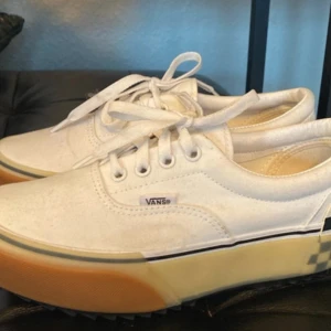 Vita sneakers från Vans - Snygga vita Vans sneakers med klassisk snörning och checkerboard-mönster på hälen. Skorna har en tjock gummisula i beige och vit, vilket ger en cool kontrast. Perfekta för en avslappnad stil.