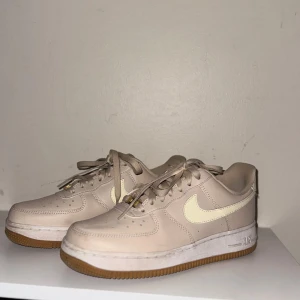 Beige Nike Air Force 1 sneakers - Snygga Nike Air Force 1 sneakers i beige skinn med klassisk vit swoosh och gummisula. Låga med snörning och gulddetaljer på skosnörena. Perfekta för dig som gillar stilrena och neutrala färger. Ikonisk siluett som alltid är trendig.